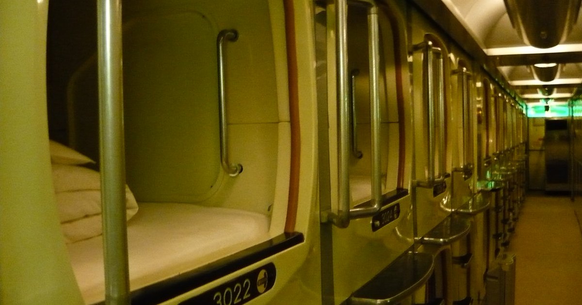Row of capsule hotel sleeping pods with bedding and numbered doors along a corridor