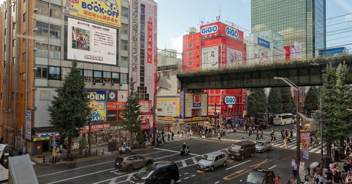 Akihabara Electric Town intersection near the station, with colorful storefront signs for electronics and game shops and a railway overpass crossing the street