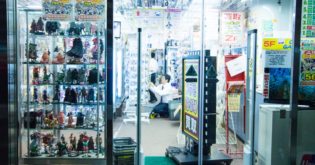 Glass display case of secondhand anime figures beside kaitori buyback signs at an Akihabara shop entrance