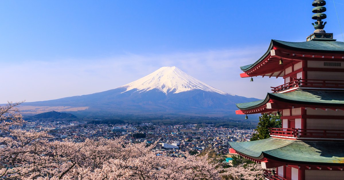 Five-story red pagoda on a hillside with cherry blossoms in front and Mt. Fuji across the town below