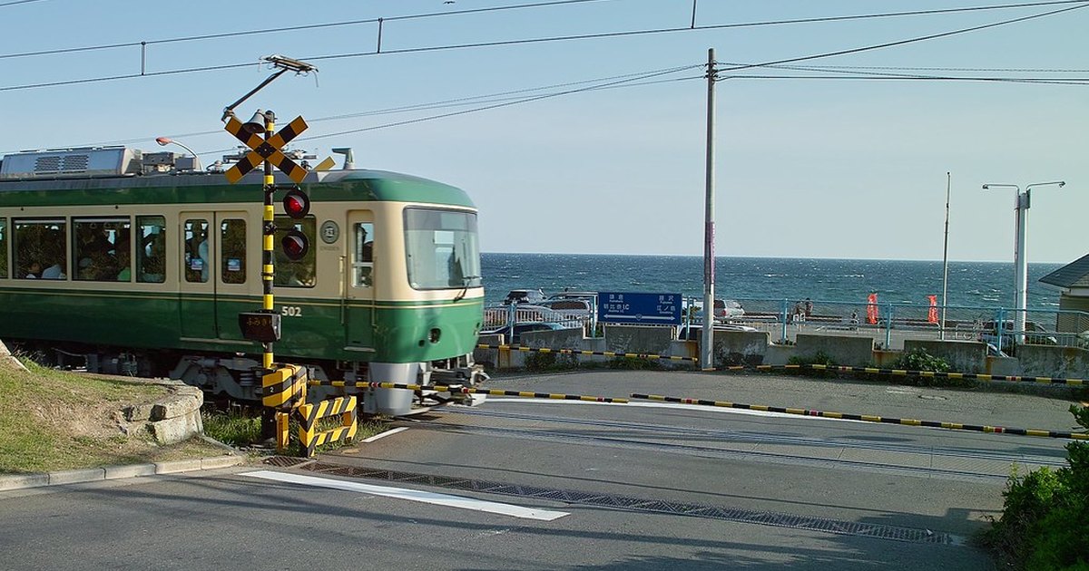 A green and cream Enoden train stopped at a level crossing beside the road, with the open sea directly behind it