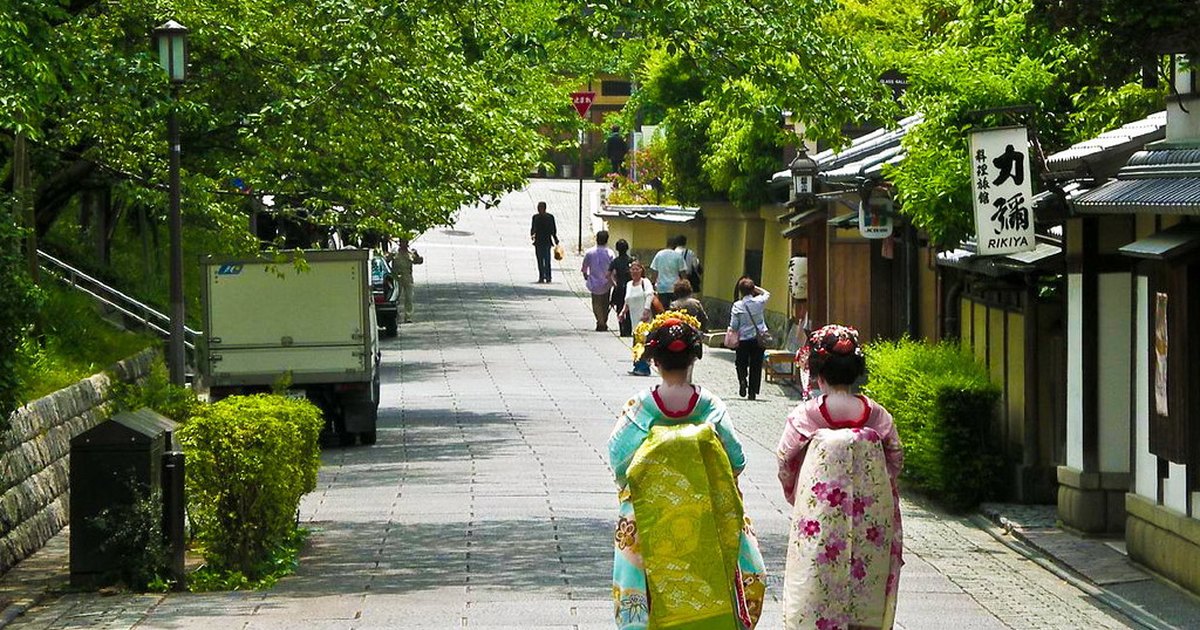 Two maiko in colorful kimono with obi sashes walking away down a stone-paved street in Kyoto's Higashiyama geisha district