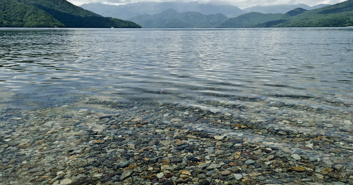 Clear shallow water over rounded pebbles at the shore of Lake Chuzenji, with forested mountains ringing the far side
