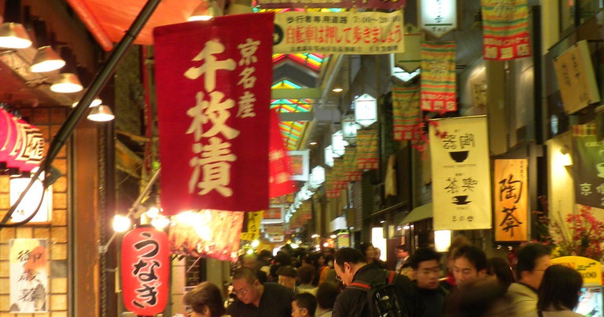 Crowded covered arcade of Nishiki Market in Kyoto, with a colorful stained-glass ceiling, hanging shop banners, and food stalls lining the narrow walkway