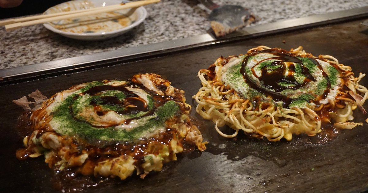 Two okonomiyaki pancakes cooking on a black teppan griddle, topped with swirls of sauce, mayonnaise and green aonori powder, one with fried noodles