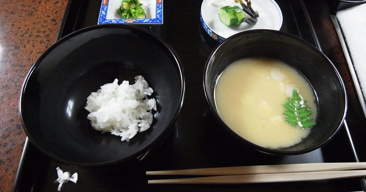 A black lacquer tray holding rice, miso soup with tofu, pickles and dressed greens with wooden chopsticks