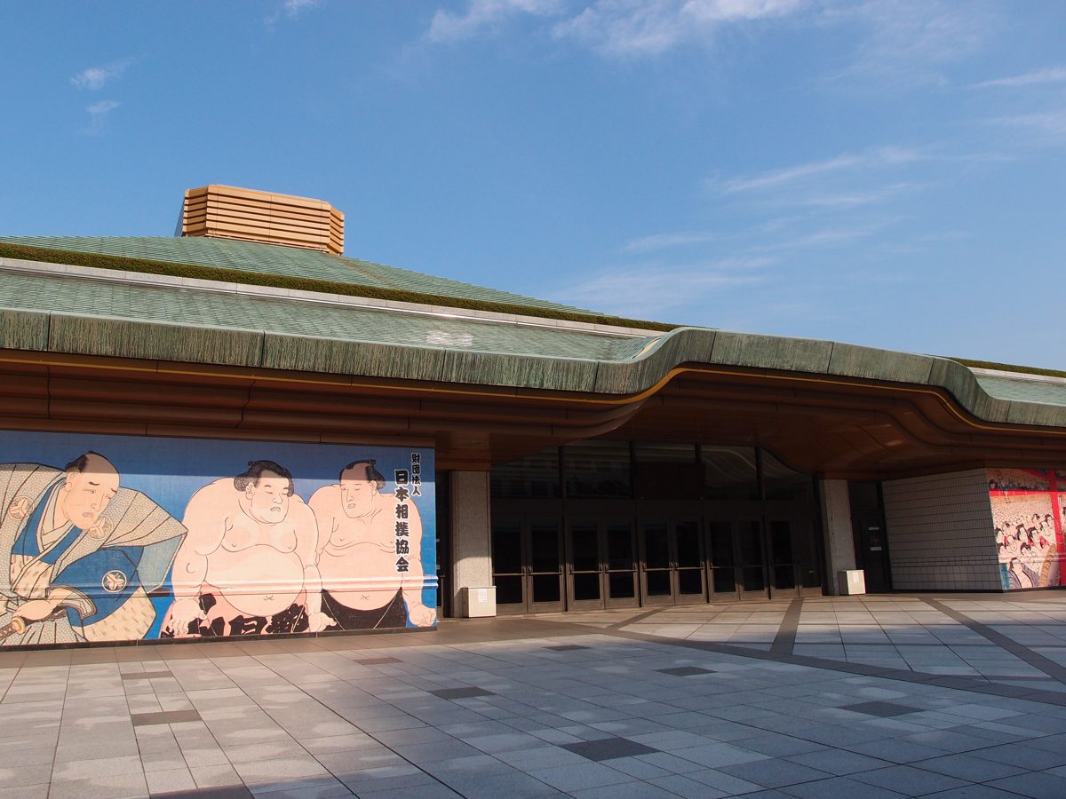 The green roof and entrance of Ryogoku Kokugikan with paintings of sumo wrestlers on the wall