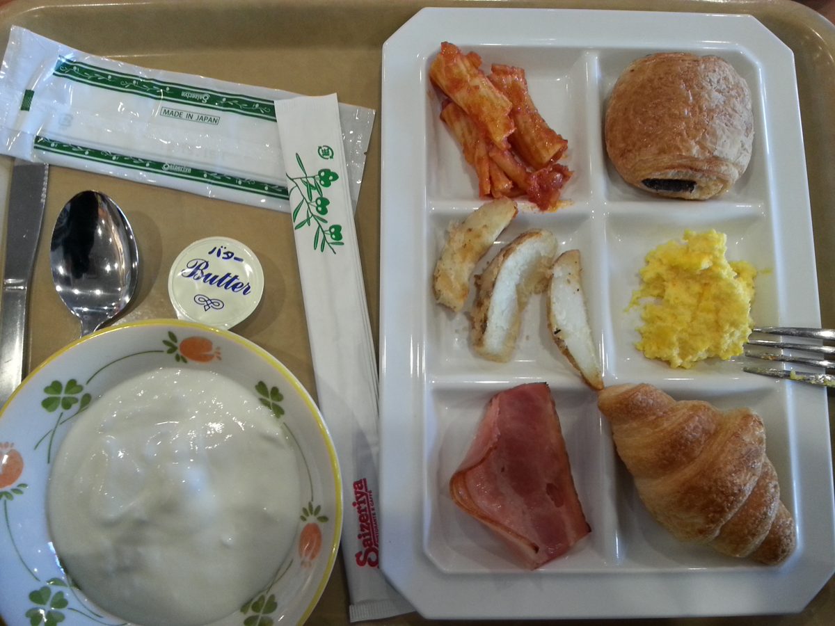 A hotel breakfast plate with a croissant, scrambled egg, ham and a bowl of yogurt