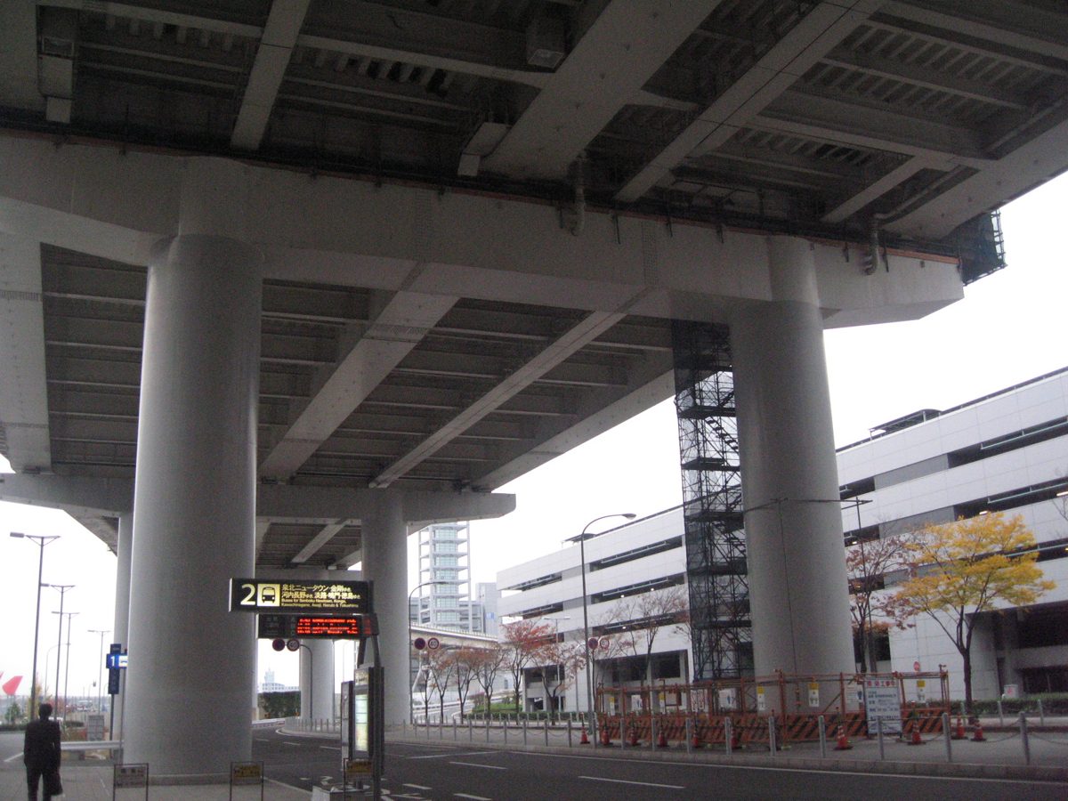 A bus stop under an elevated road at Kansai International Airport, with a multi-storey building on the right