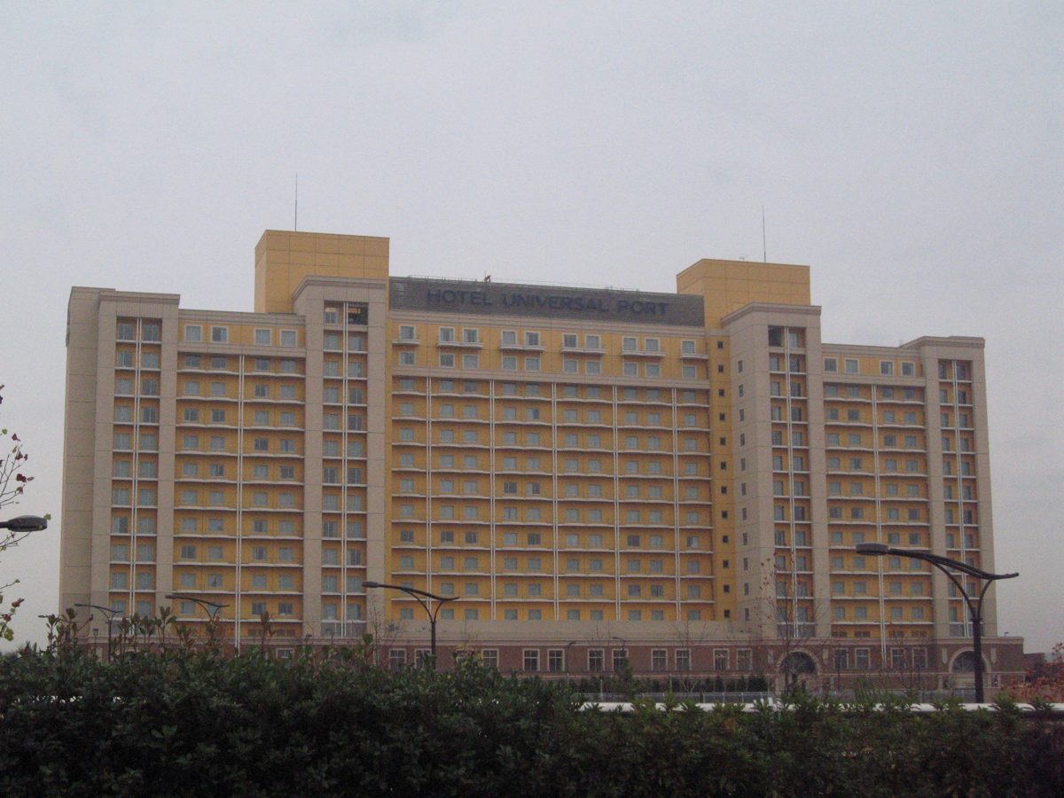 Hotel Universal Port, a wide cream building, behind trees and street lamps on a cloudy day