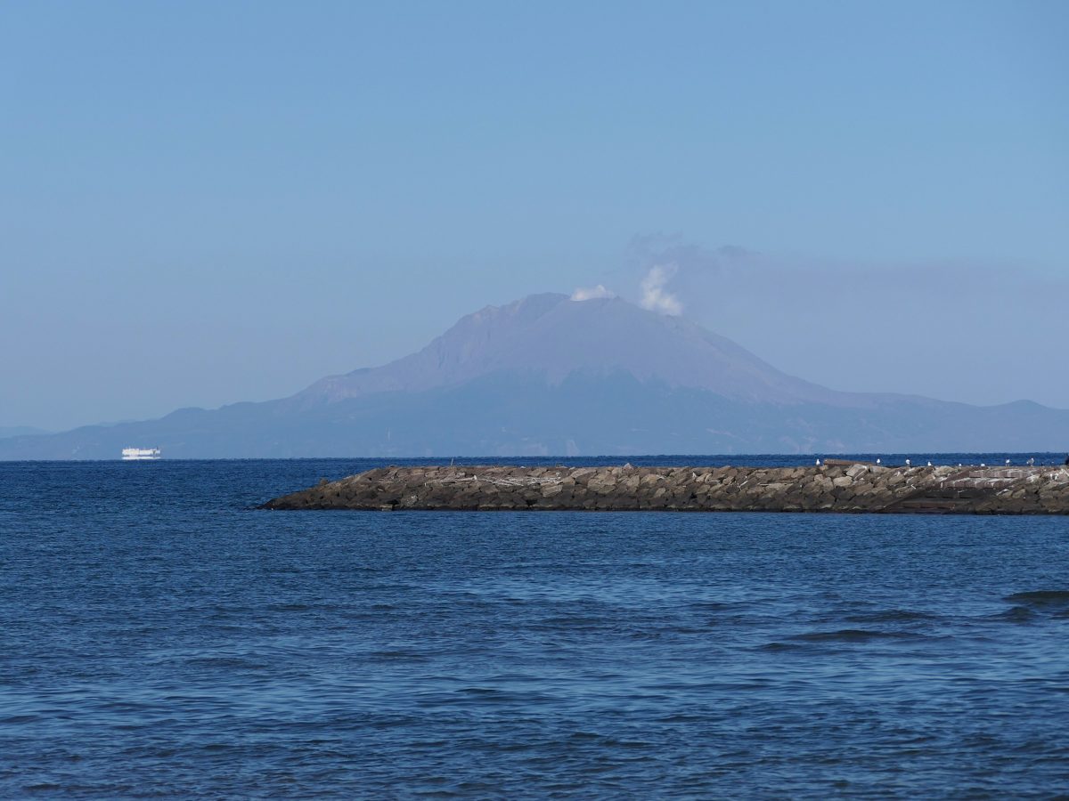 A smoking volcano across calm blue water, seen from a beach