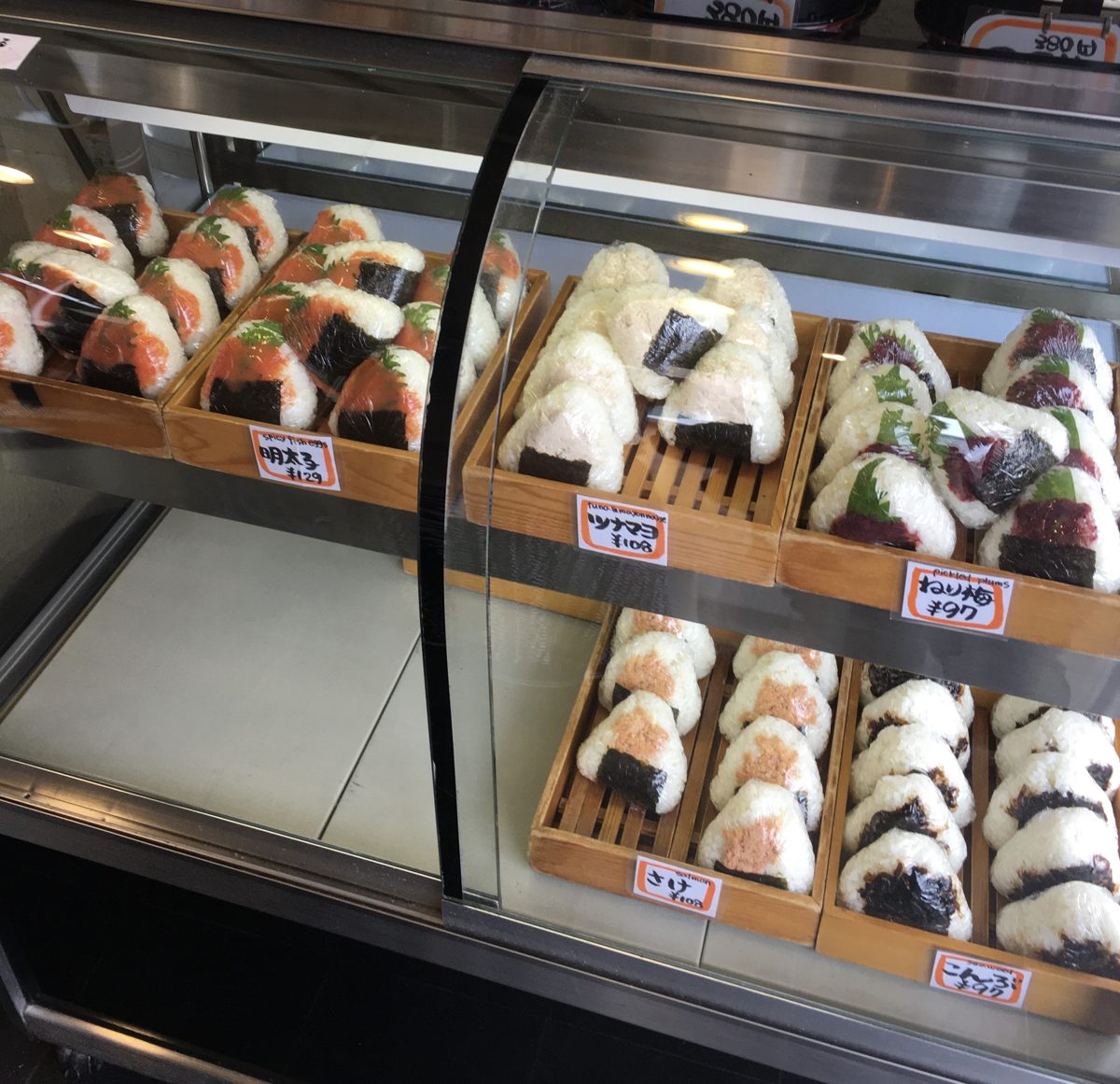 Rows of onigiri in wooden trays with handwritten price tags in a glass display case