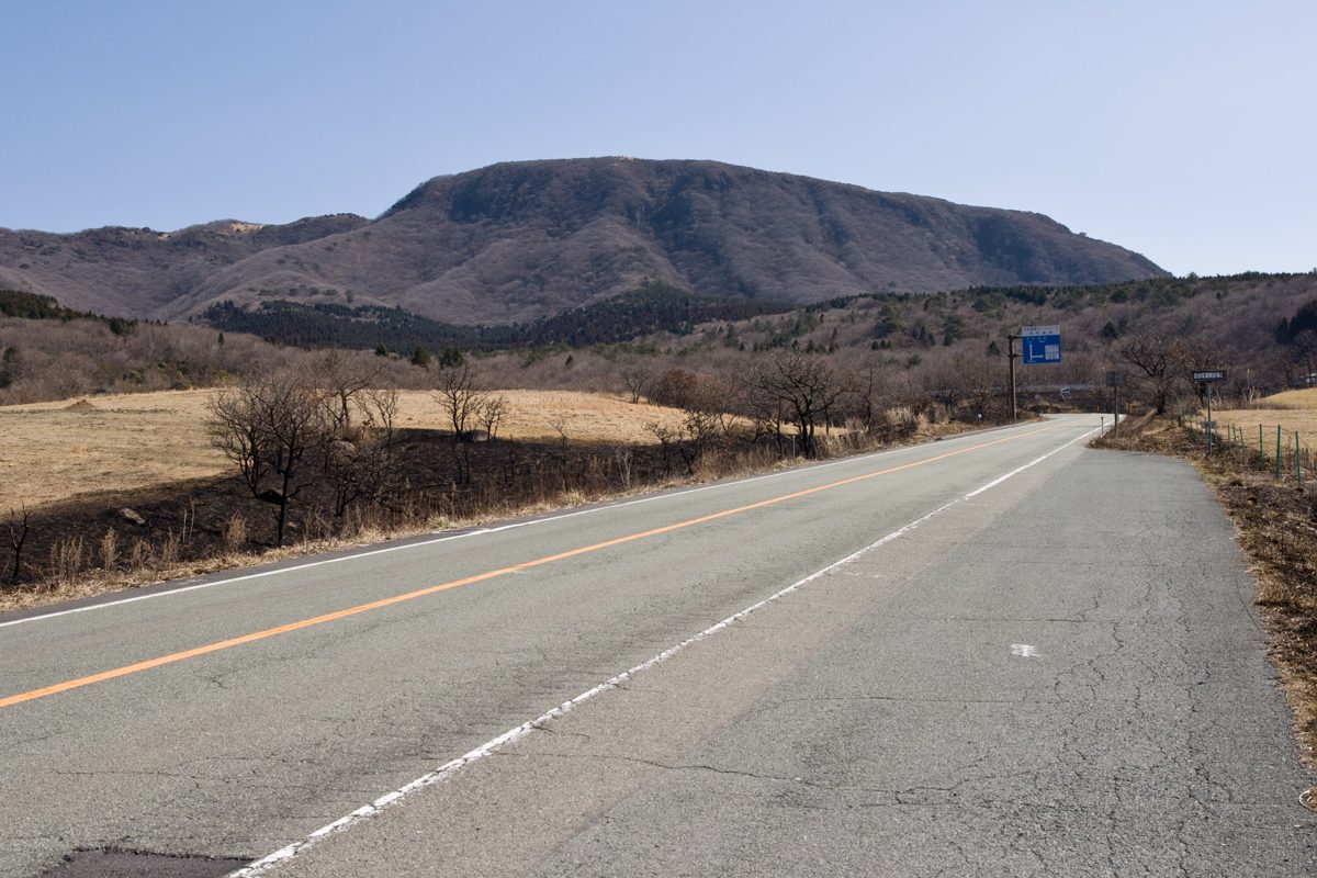 An empty two-lane road across dry highland grass toward a mountain