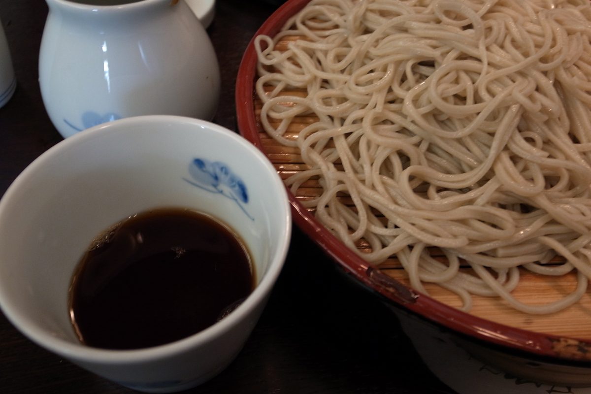 A red-rimmed basket of cold soba with a cup of dipping sauce and a small jug