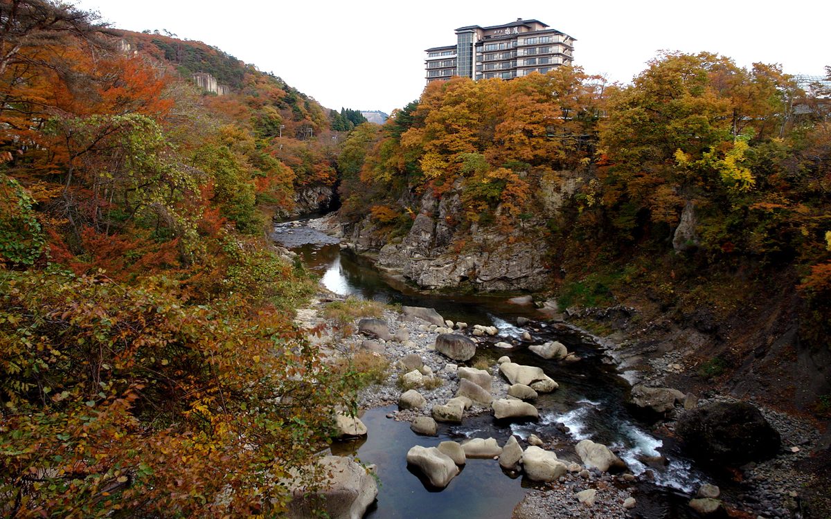 Rocky river gorge with autumn leaves and a tall hotel building on the far bank at Akiu Onsen