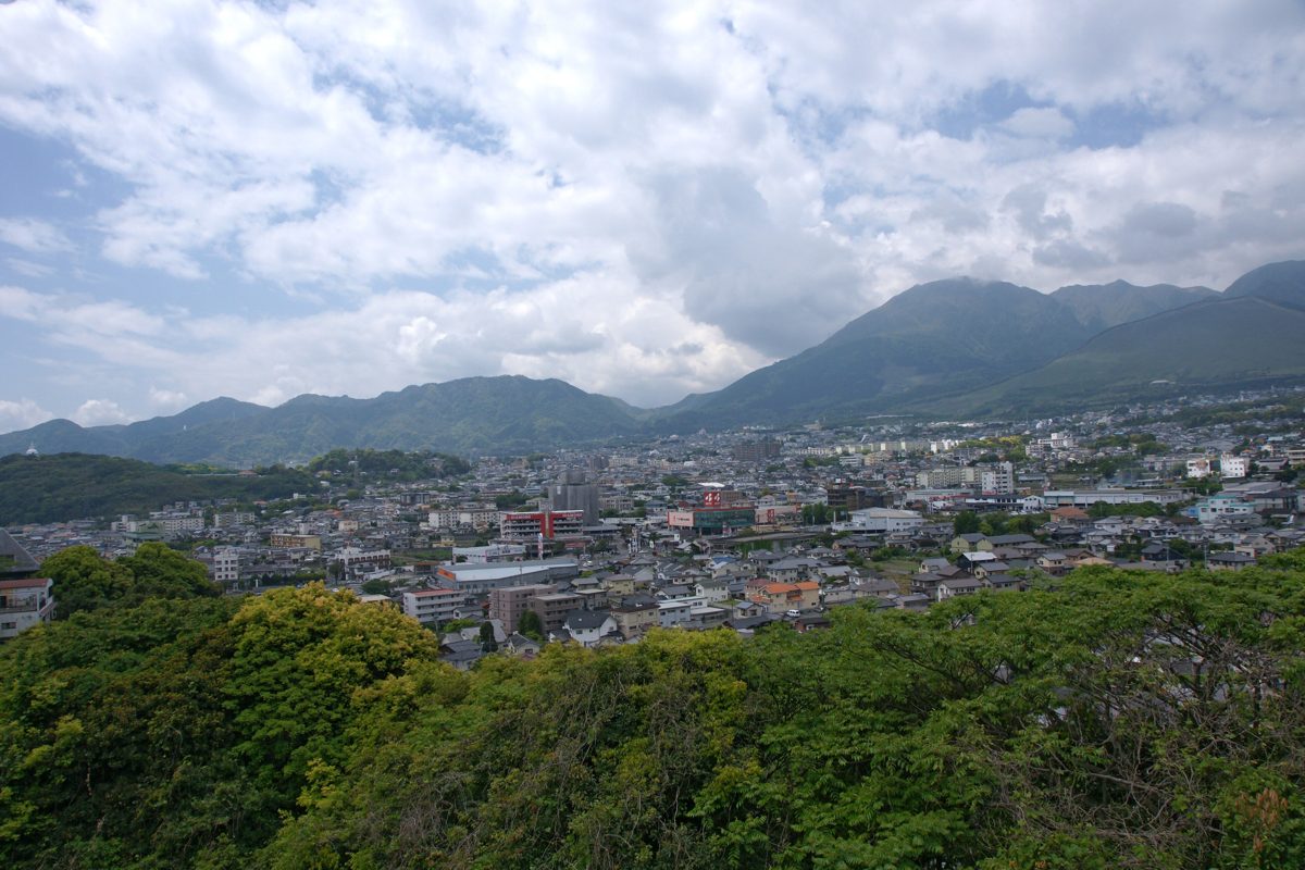 Rooftops of Beppu below green mountains under a partly cloudy sky, seen from a hillside