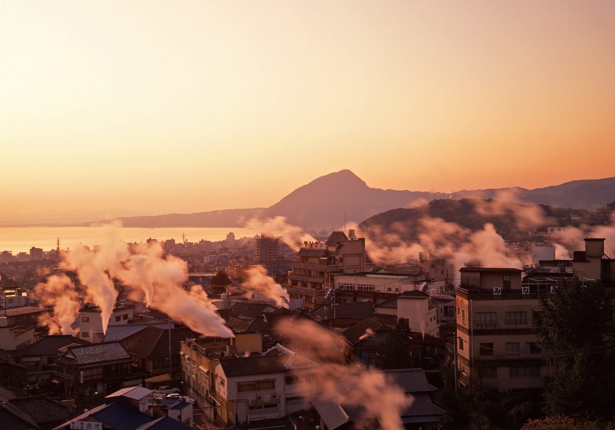Steam from hot springs rising over Beppu rooftops at sunset with a mountain behind