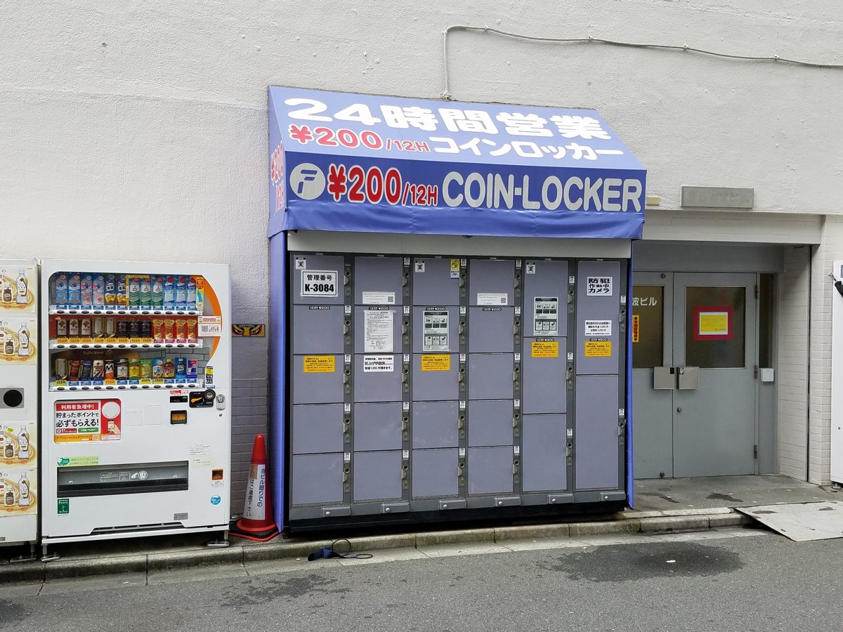 A bank of blue-gray coin lockers with a sign for 200-yen, 24-hour lockers, next to a drink vending machine