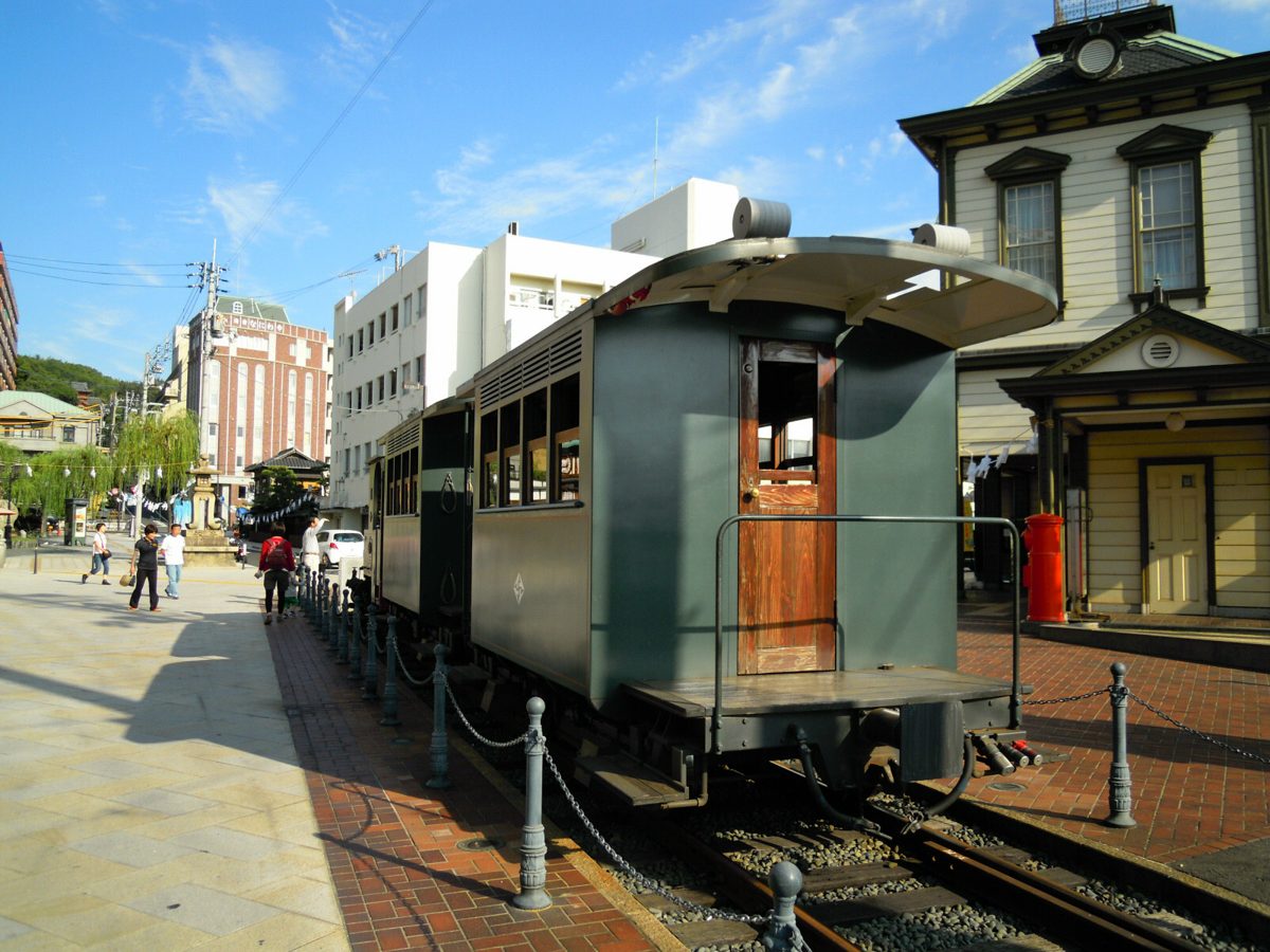 A green steam-style Botchan Train car standing on tracks beside a paved plaza on a sunny day