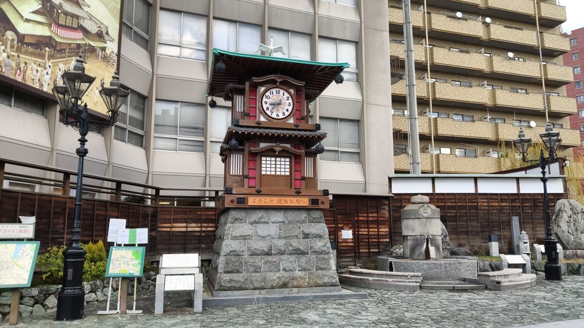 A wooden Japanese-style clock tower on a stone base, with a small fountain and street lamps in front of tall buildings