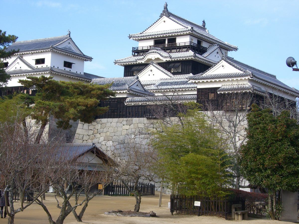 The white main and small towers of Matsuyama Castle above a stone wall under a blue sky