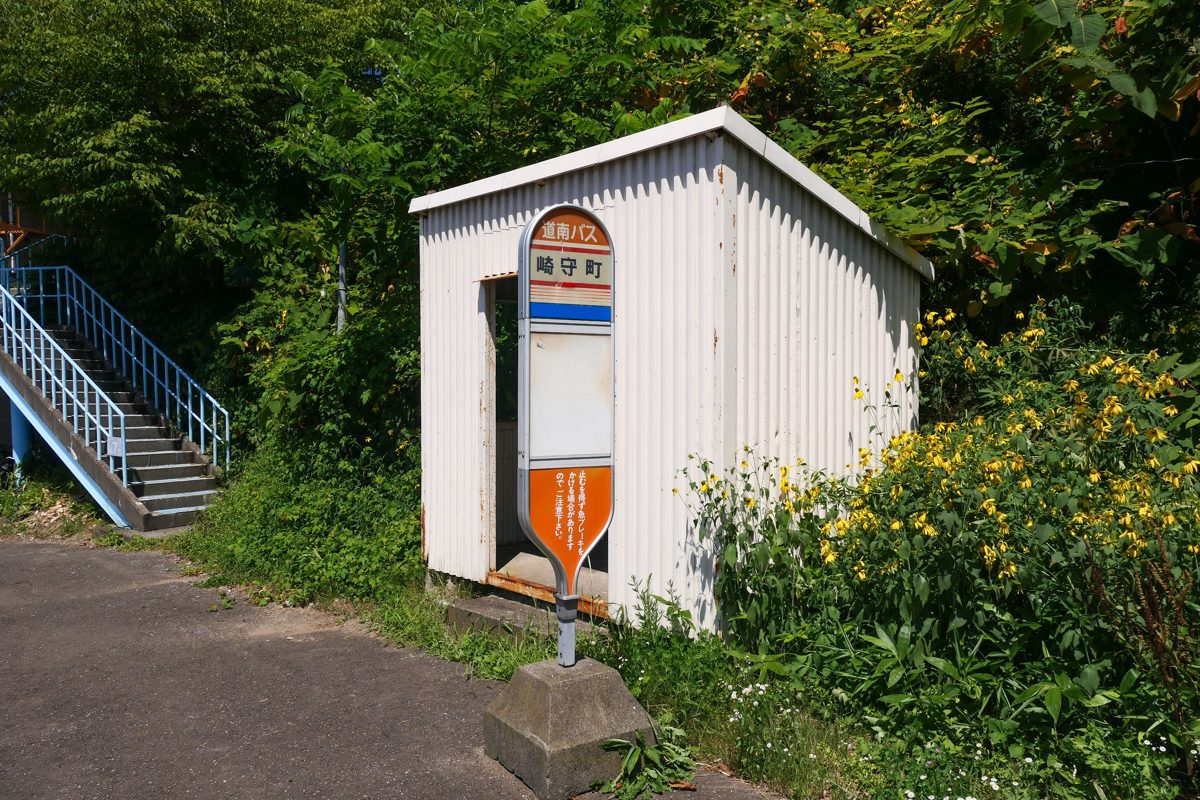 A small white bus shelter with a stop sign beside green bushes