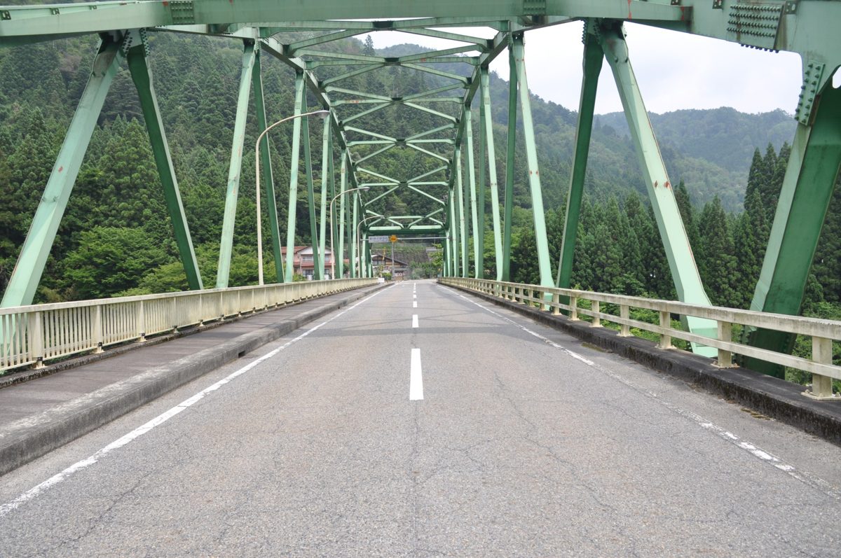A green steel truss bridge carrying a two-lane road between wooded hills on a clear day