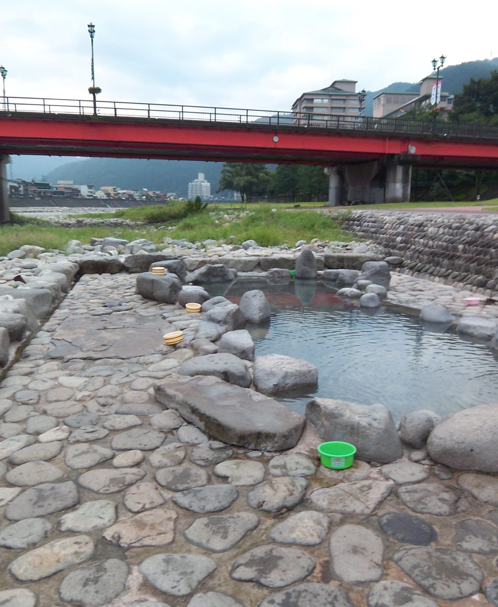 A stone-lined outdoor hot-spring pool on a riverbank, with a red road bridge and hills behind it