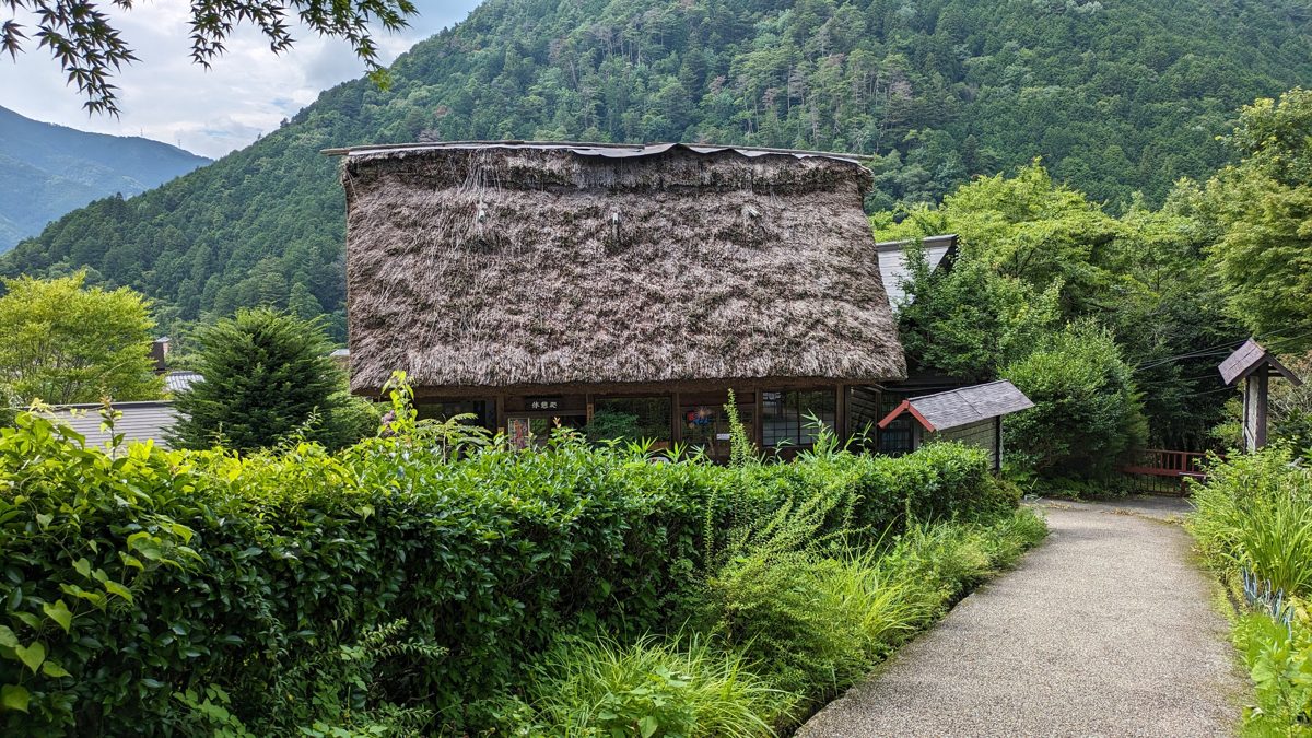A thatched-roof farmhouse beside a garden path, with forested hills behind