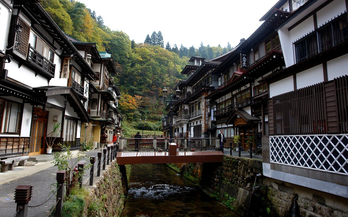 Wooden multi-story inns on both sides of a small river with a footbridge, autumn trees on the hillside behind