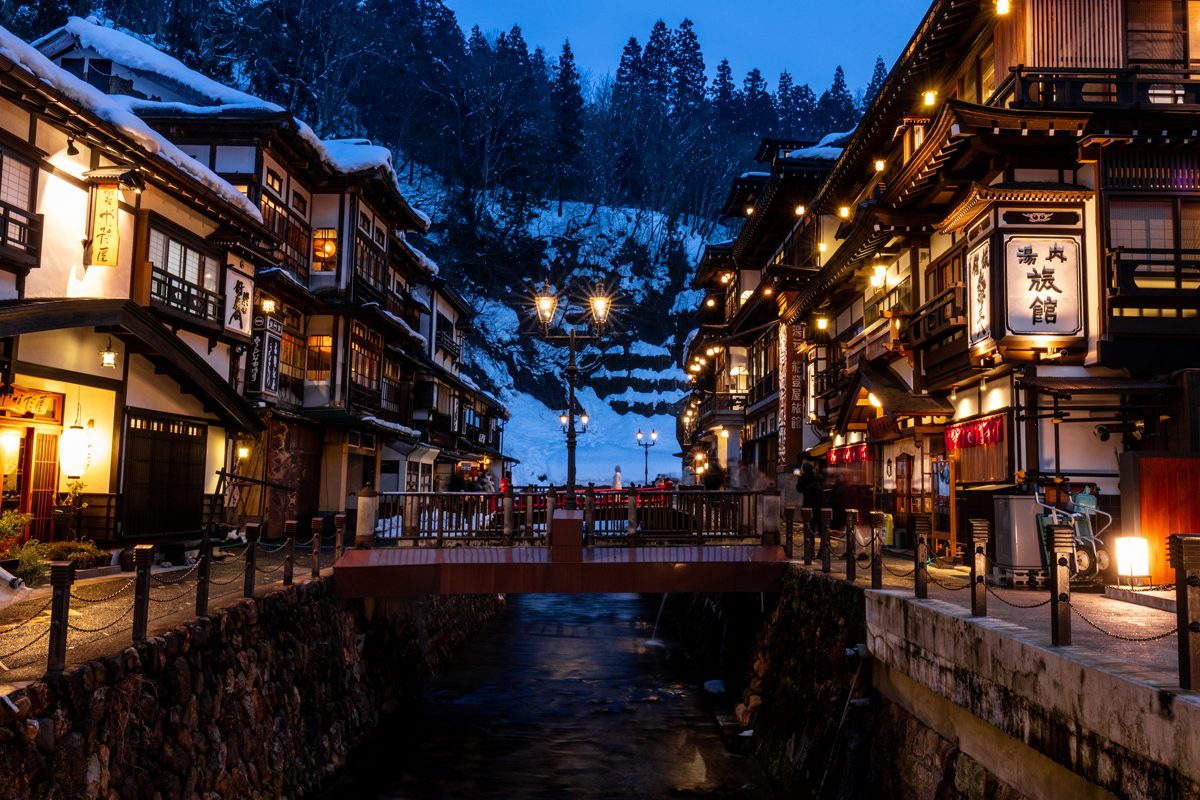 Lit wooden inns along a river at dusk with snow on the hills