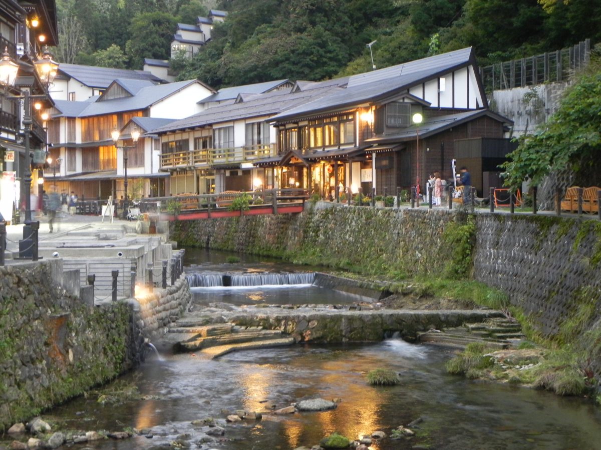 The Ginzan River with a small weir, stone banks and wooden buildings with lit windows at dusk