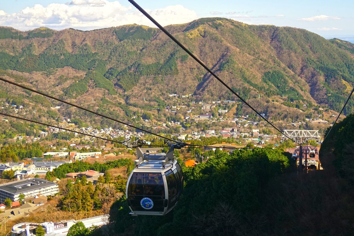 A ropeway gondola over a green hillside with a town and mountains behind, Hakone