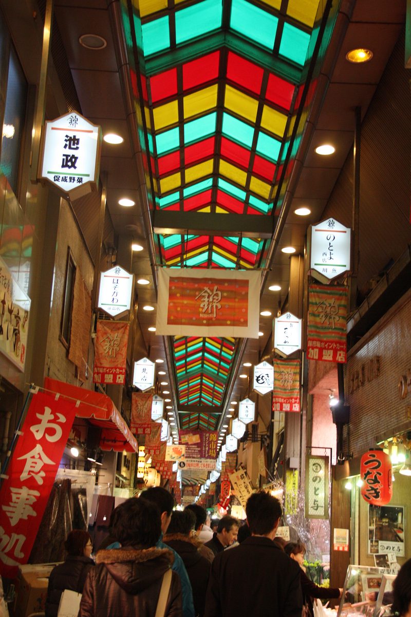 Covered shopping arcade with a colorful stained-glass roof, hanging shop signs, and shoppers