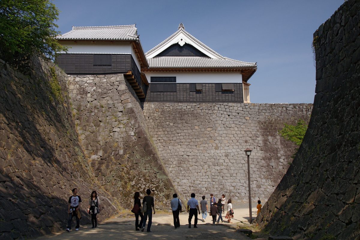 Visitors walking between the tall curved stone walls of Kumamoto Castle with turrets above