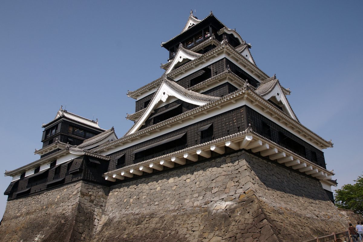 The main keep of Kumamoto Castle with black walls and white gables above a curved stone base, under a blue sky