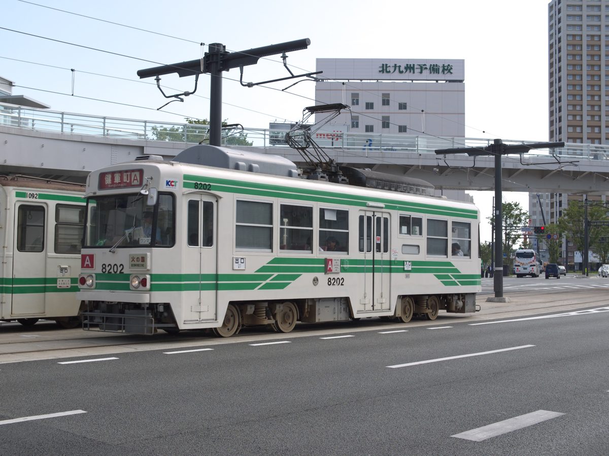 A green and white Kumamoto City Tram 8200 series streetcar on a wide city street with overhead wires