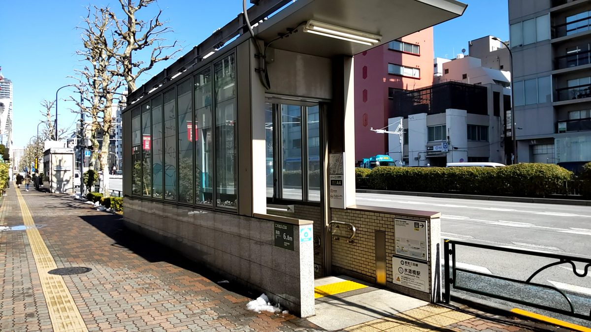A glass-walled Toei Mita Line subway entrance on a sidewalk near Suidobashi