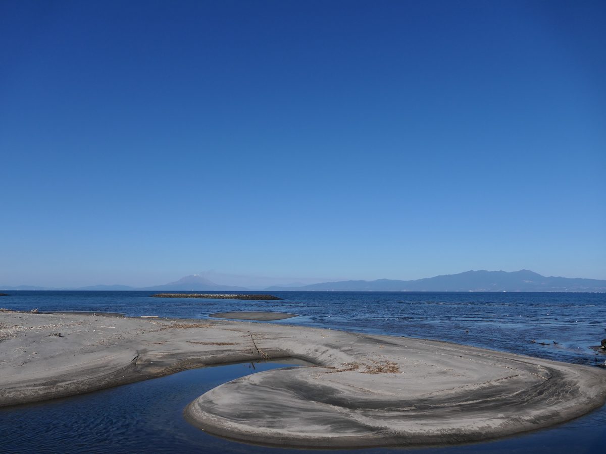 A sandy point on a calm sea under a clear blue sky, with low mountains on the horizon
