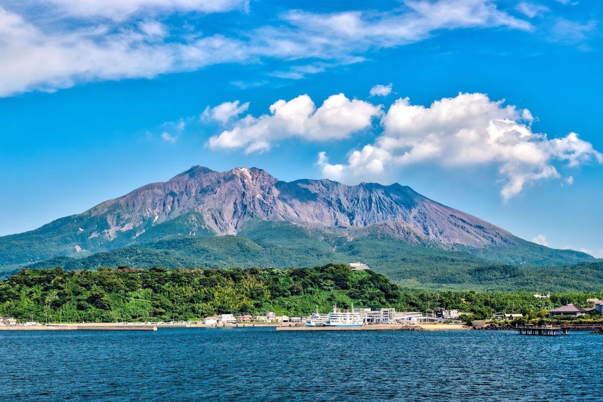 Sakurajima volcano under white clouds, with a green shoreline and a harbor across blue water