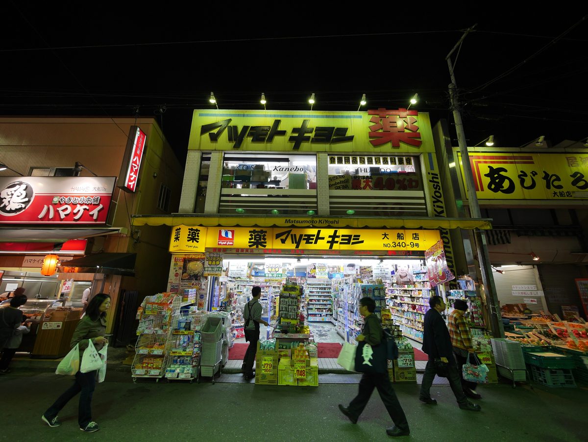 A brightly lit Matsumoto Kiyoshi drugstore at night with shelves of goods at the open entrance and people walking past