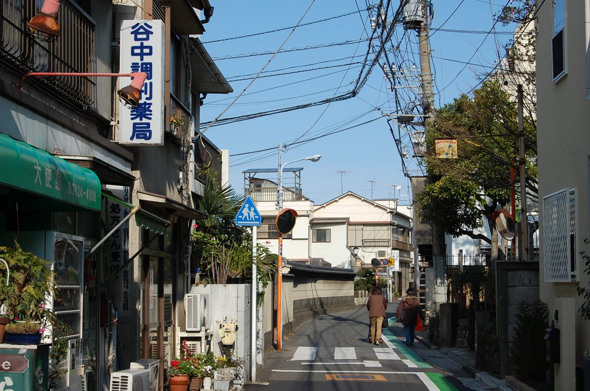 A narrow residential street with a vertical blue and white sign for Yanaka Chozai Yakkyoku, a dispensing pharmacy