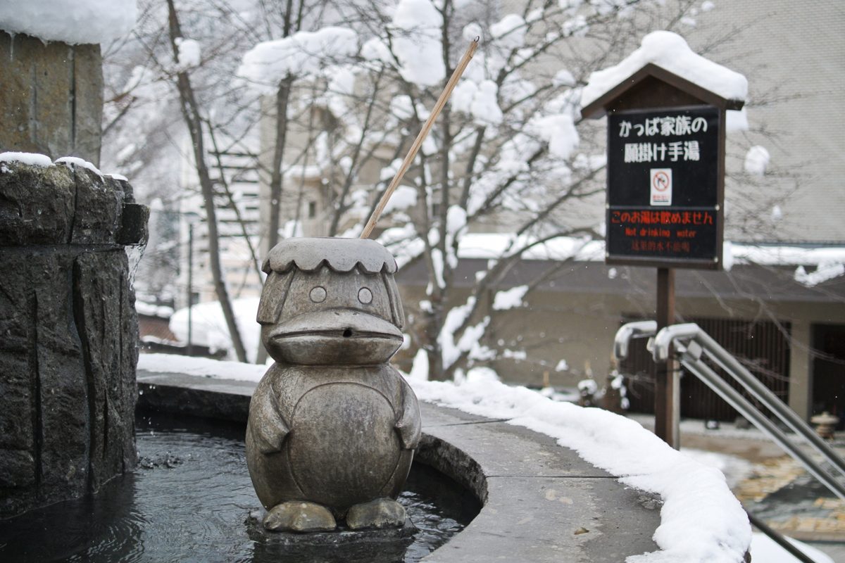 A stone kappa statue beside a small hot-spring hand bath in the snow