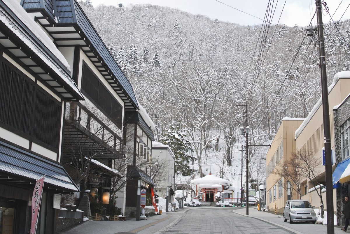 A snowy street lined with low buildings in Jozankei Onsen, with mountains behind