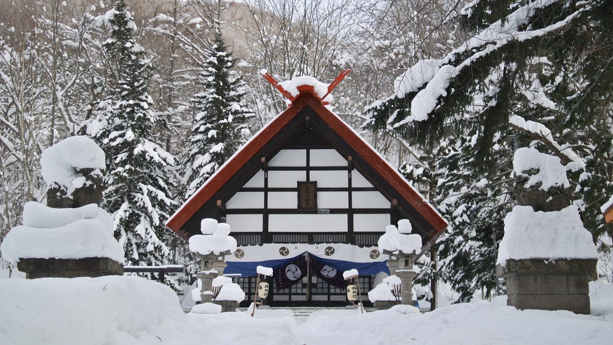 A shrine building with a red roof and stone guardian statues covered in snow, surrounded by snowy trees