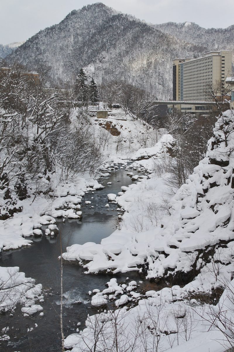 A river running through a snowy valley with forested mountains and a large building in the distance