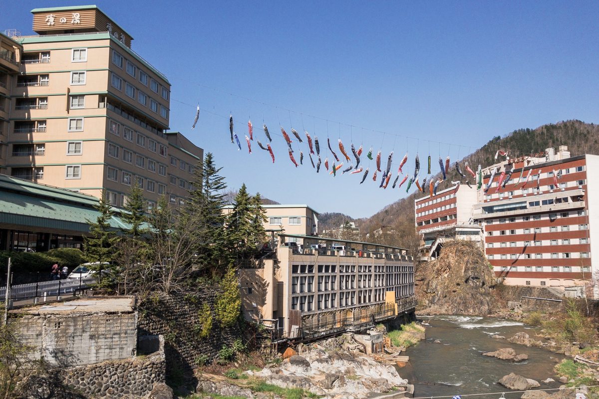 Hotels on both banks of a rocky river gorge at Jozankei Onsen under a blue sky, with carp streamers strung across the river