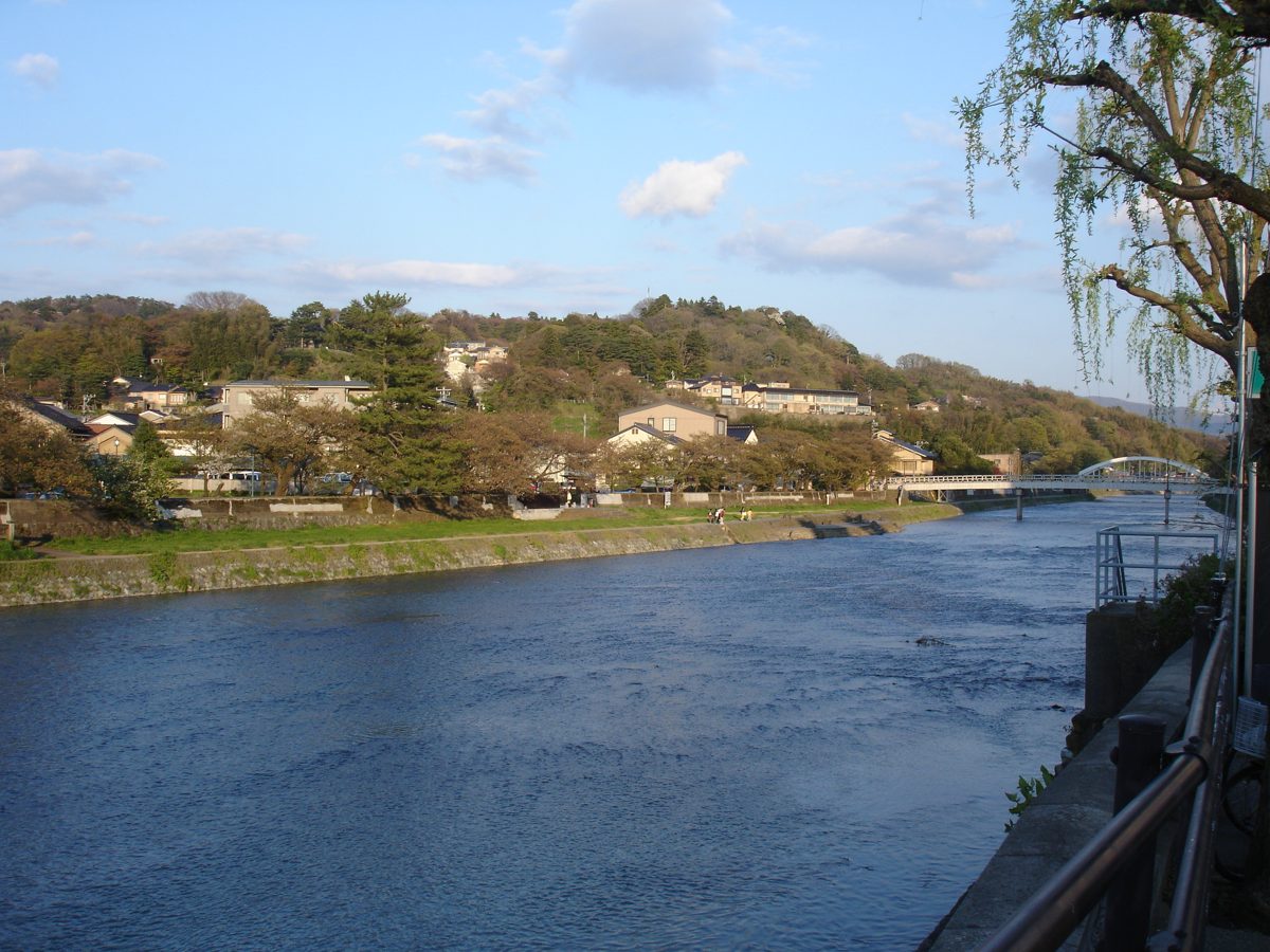 A wide river with houses and a wooded hill on the far bank and a footbridge in the distance under a blue sky