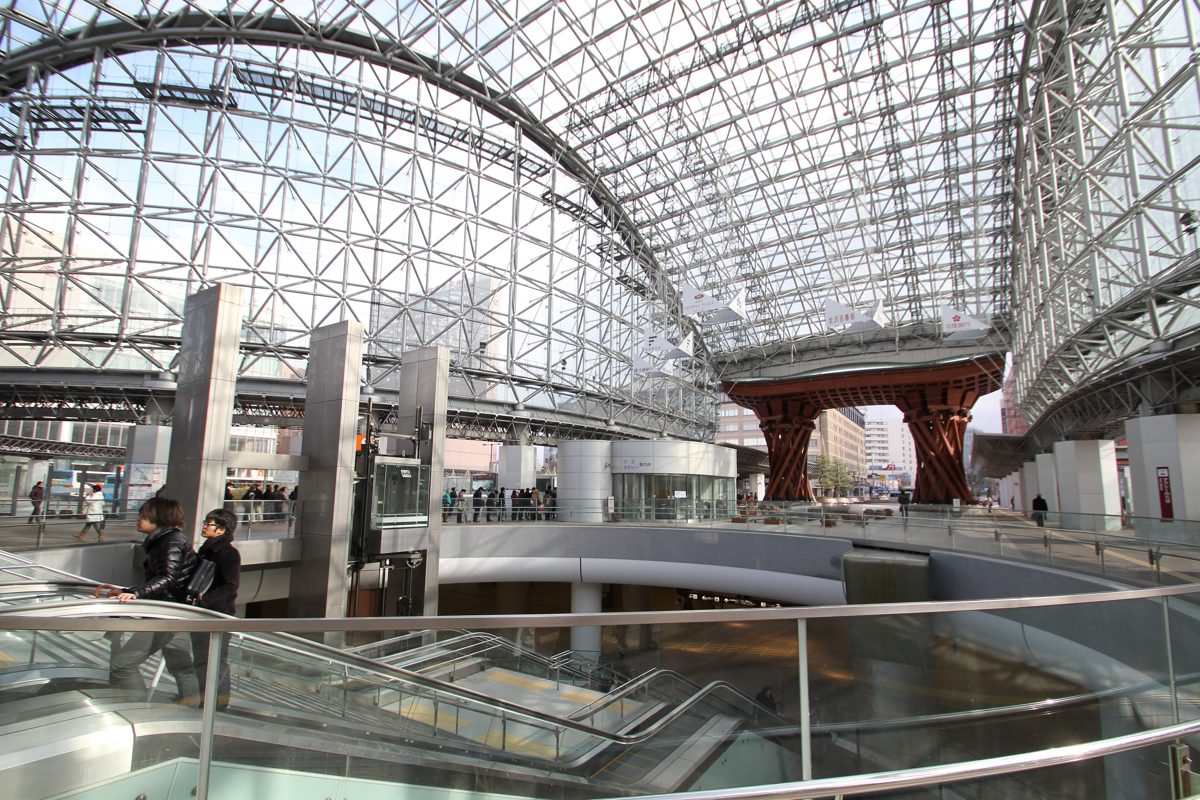 Glass and steel dome roof inside Kanazawa Station, with the red wooden Tsuzumi-mon gate beyond