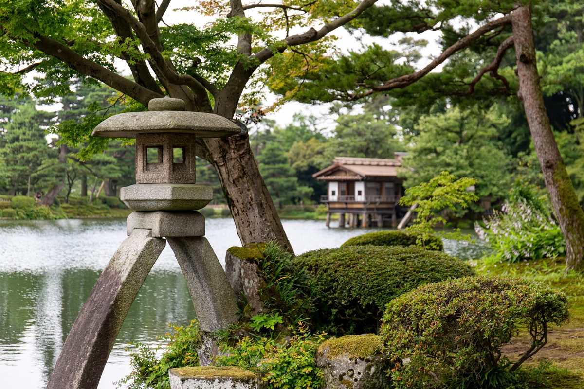 A two-legged stone lantern beside a pond with a wooden teahouse and pine trees at Kenroku-en garden